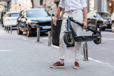 cropped view of tattooed middle aged man in white shirt carrying electric scooter on street 