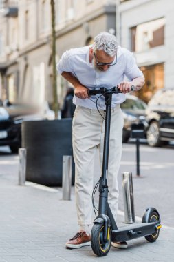 full length of mature man in glasses looking at electric scooter on street  clipart