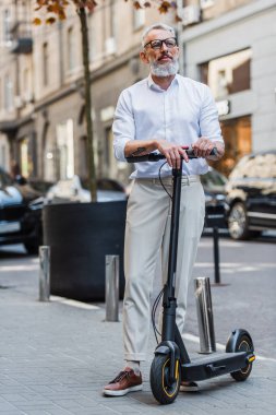 full length of middle aged man in glasses standing with electric scooter on street 