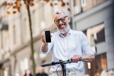 happy middle aged man in glasses holding cellphone with blank screen and standing near e-scooter 