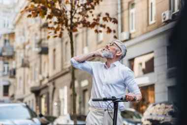 happy middle aged man in glasses talking on smartphone and looking up while standing near e-scooter 