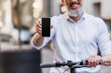 cropped view of happy middle aged man holding cellphone with blank screen and standing near e-scooter 