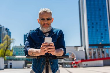 mature man in sunglasses using cellphone near electric scooter on urban street