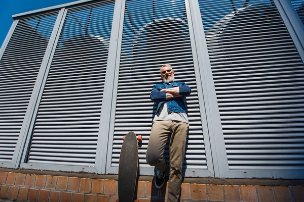 low angle view of positive and middle aged man standing with crossed arms near longboard on street