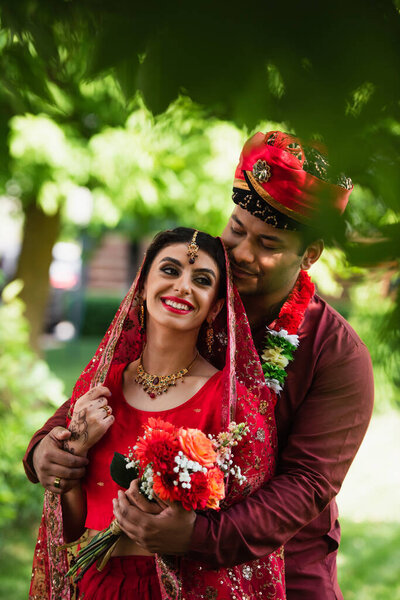 happy indian man hugging cheerful bride in headscarf and sari outdoors