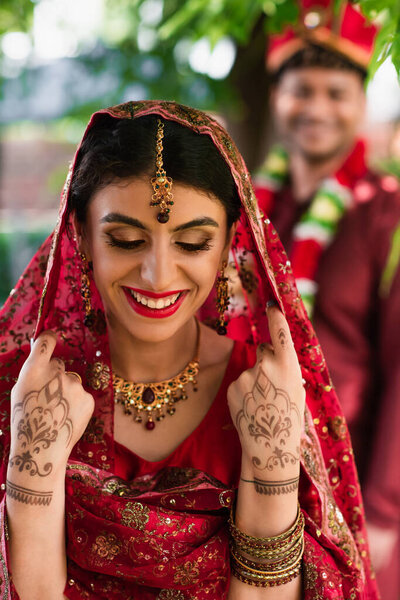 cheerful indian bride in sari and headscarf near blurred man in turban on background 