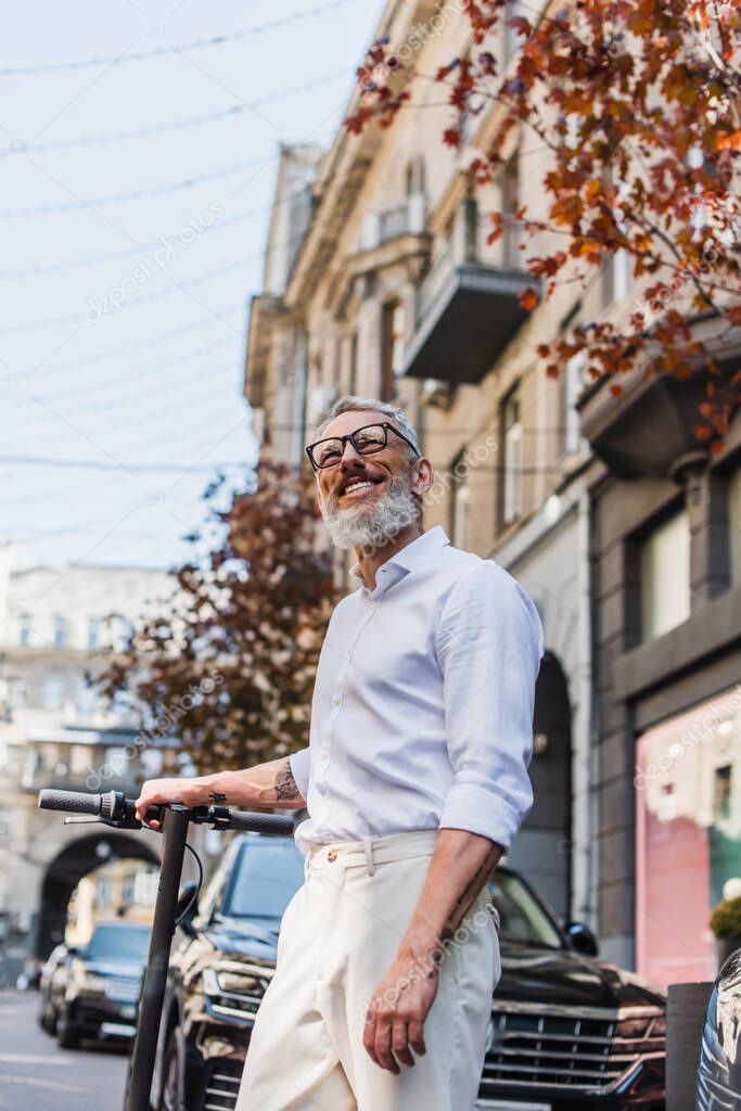 low angle view of joyful middle aged man in white shirt looking up near electric scooter on street 