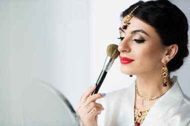 Mehndi ile Hintli gelin beyaz aynanın yanında makyaj fırçasıyla yüz pudrası sürüyor. 
