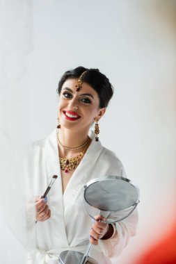 Neşeli Hintli gelin, elinde ayna ve beyaz kozmetik fırçasıyla 