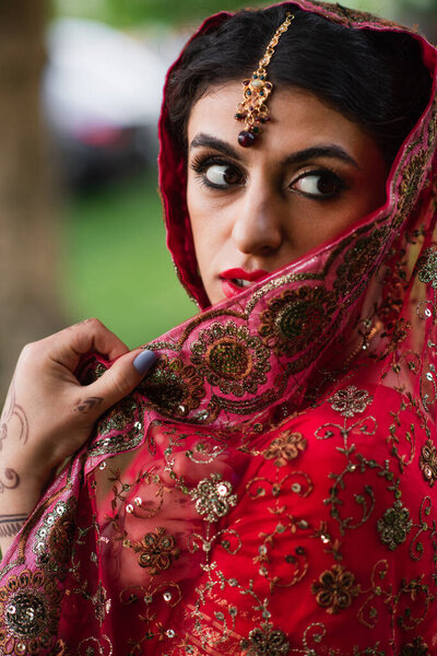 young indian bride in red sari adjusting headscarf with ornament 