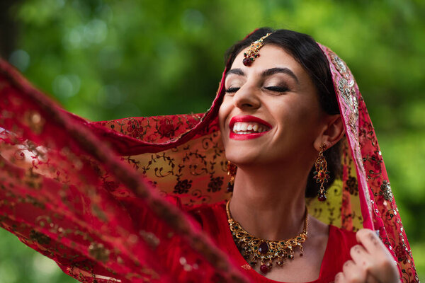 positive indian bride in red sari adjusting headscarf with ornament 