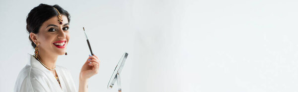счастливая индийская невеста с mehndi держа зеркало и косметической щеткой на белом, баннер