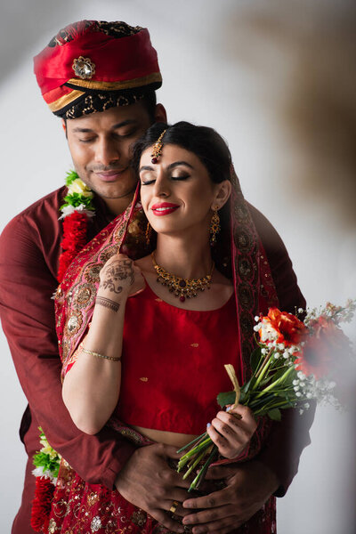 pleased indian man hugging happy bride with bouquet of flowers on white