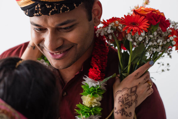 happy indian man in turban looking at blurred bride with mehndi holding flowers isolated on white