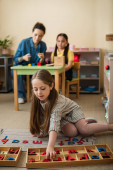 kid on floor playing with wooden letters near teacher and asian girl on blurred background