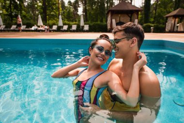 Güneş gözlüklü gülümseyen adam yüzme havuzunda gözleri kapalı kız arkadaşına sarılıyor. 