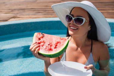 Smiling woman holding watermelon and plate in blurred swimming pool  clipart