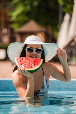Karpuz ve güneş şapkasını havuzda tutarken gülümseyen genç bir kadın. 