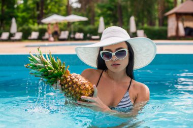 Yüzme havuzunda su damlalarıyla ananas tutan genç bir kadın. 