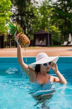 Güneş gözlüklü kadın yüzme havuzunda ananas yükseliyor. 