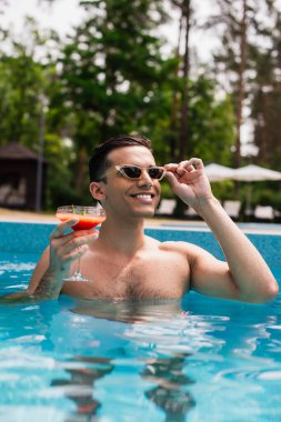 Havuzda güneş gözlüğü tutan neşeli bir adam. 