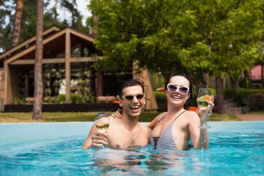Young couple holding glasses of white wine in swimming pool  clipart