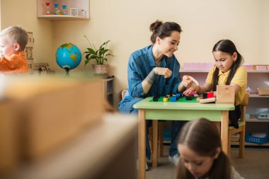 Öğretmen bulanık ön planda Asyalı bir kızla oynarken çok renkli küpleri gösteriyor.