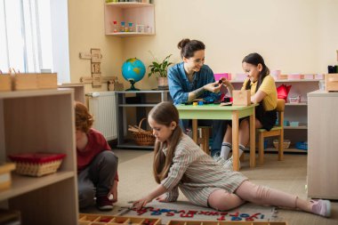 Yerdeki çocuklar öğretmenlerinin yanında ahşap harflerle oynuyorlar ve arka planda çok renkli küpleri olan bir kız var.