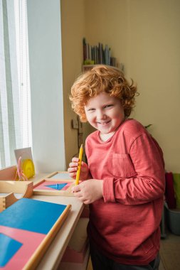 Montessori okulunda elinde renkli kalemle kameraya bakan neşeli, kızıl çocuk.