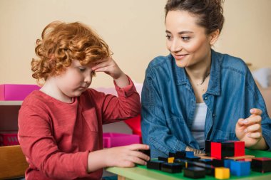 Öğretmen, Montessori okulunda düşünceli bir çocuğun ve renkli küplerin yanında gülümsüyor.