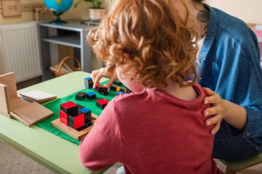 Montessori okulunda öğretmenle eğitim oyunu oynayan kızıl saçlı çocuğun arka görünüşü