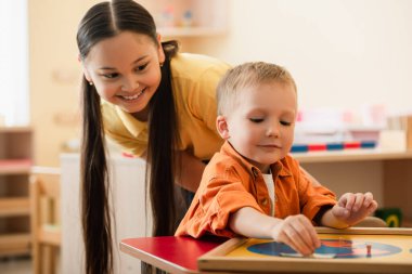gülümseyen Asyalı kız Montessori okulunda haritayı birleştiren çocuğa bakıyor.