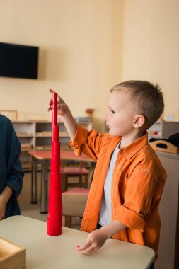 Çocuk Montessori okulunda öğretmenin yanında kırmızı ahşap silindirlerden kule yapıyor.