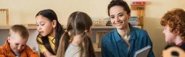 Montessori okulunda çocuklar arasında gülümseyen mutlu genç öğretmen, afiş