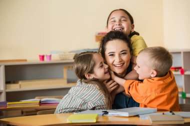 overjoyed multiethnic kids embracing happy teacher in montessori school clipart