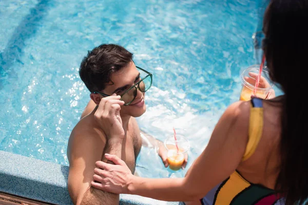 Overhead view of man holding sunglasses near blurred girlfriend with drink on poolside