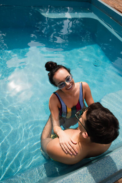 Вид сверху на улыбающуюся женщину, отдыхающую с парнем в солнечных очках в бассейне 