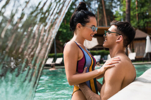 Side view of young man hugging pretty girlfriend near fountain in swimming pool 