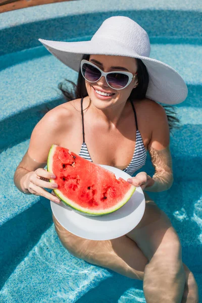 High angle view of positive woman in swimwear holding watermelon in swimming pool