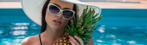 戴着太阳镜的年轻女子拿着菠萝靠近模糊的游泳池 — 图库照片