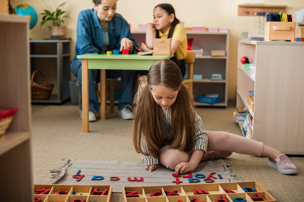 girl on floor playing with wooden letters near asian girl and teacher on blurred background