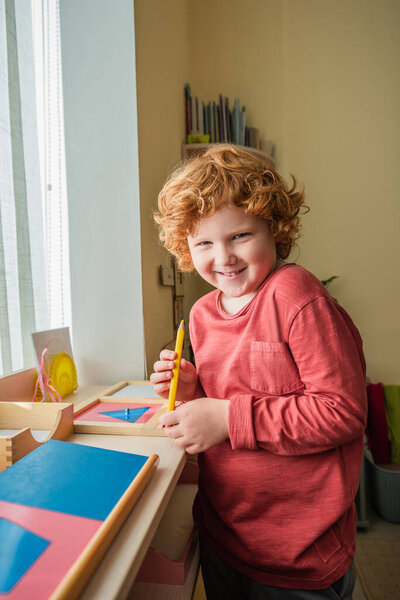 cheerful, redhead boy looking at camera while holding color pencil in montessori school