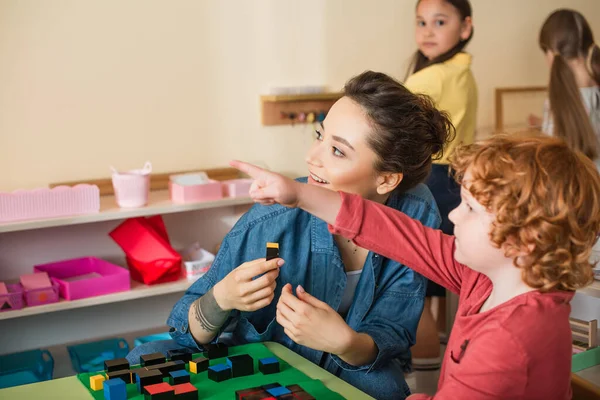 Rothaariger Junge Zeigt Mit Finger Auf Lächelnden Lehrer Mit Holzwürfel — Stockfoto