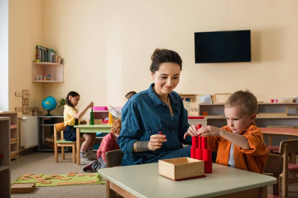 Tânăr Profesor Zâmbind Lângă Băiat Făcând Turn Din Cilindri Lemn — Fotografie, imagine de stoc