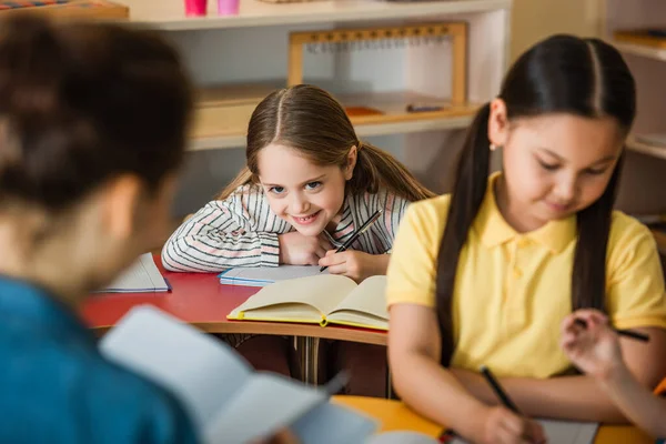 Fröhliches Kind Schreibt Während Des Unterrichts Notizbuch Neben Verschwommenem Lehrer — Stockfoto