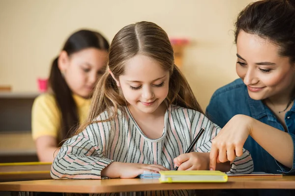 Professor Apontando Perto Sorrindo Criança Escrevendo Notebook Menina Asiática Fundo — Fotografia de Stock