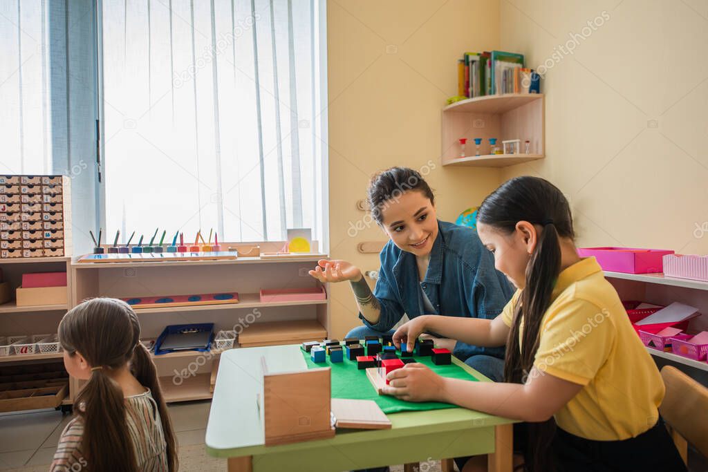 smiling teacher gesturing while playing multicolored cubes game with asian girl