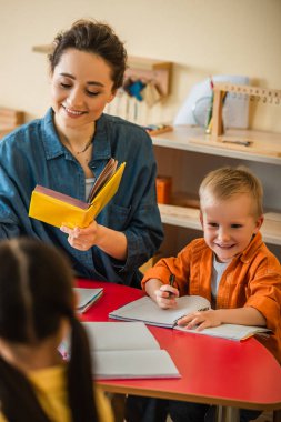 Gülümseyen öğretmen mutlu çocuğun yanında kitap tutuyor ve Montessori okulunda kızı bulanıklaştırıyor.