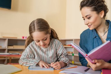 Montessori okulunda dikte yazan konsantre kızın yanında gülümseyen mutlu öğretmen.