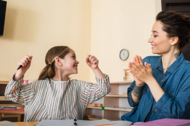 Mutlu öğretmen Montessori okulunda zafer jesti gösteren heyecanlı çocuğu alkışlıyor.